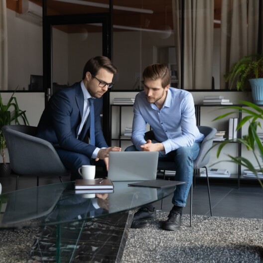 Colleagues using laptop, discussing strategy, sharing ideas, working on project in modern office together, serious mentor coach wearing glasses training new employee intern, explaining task