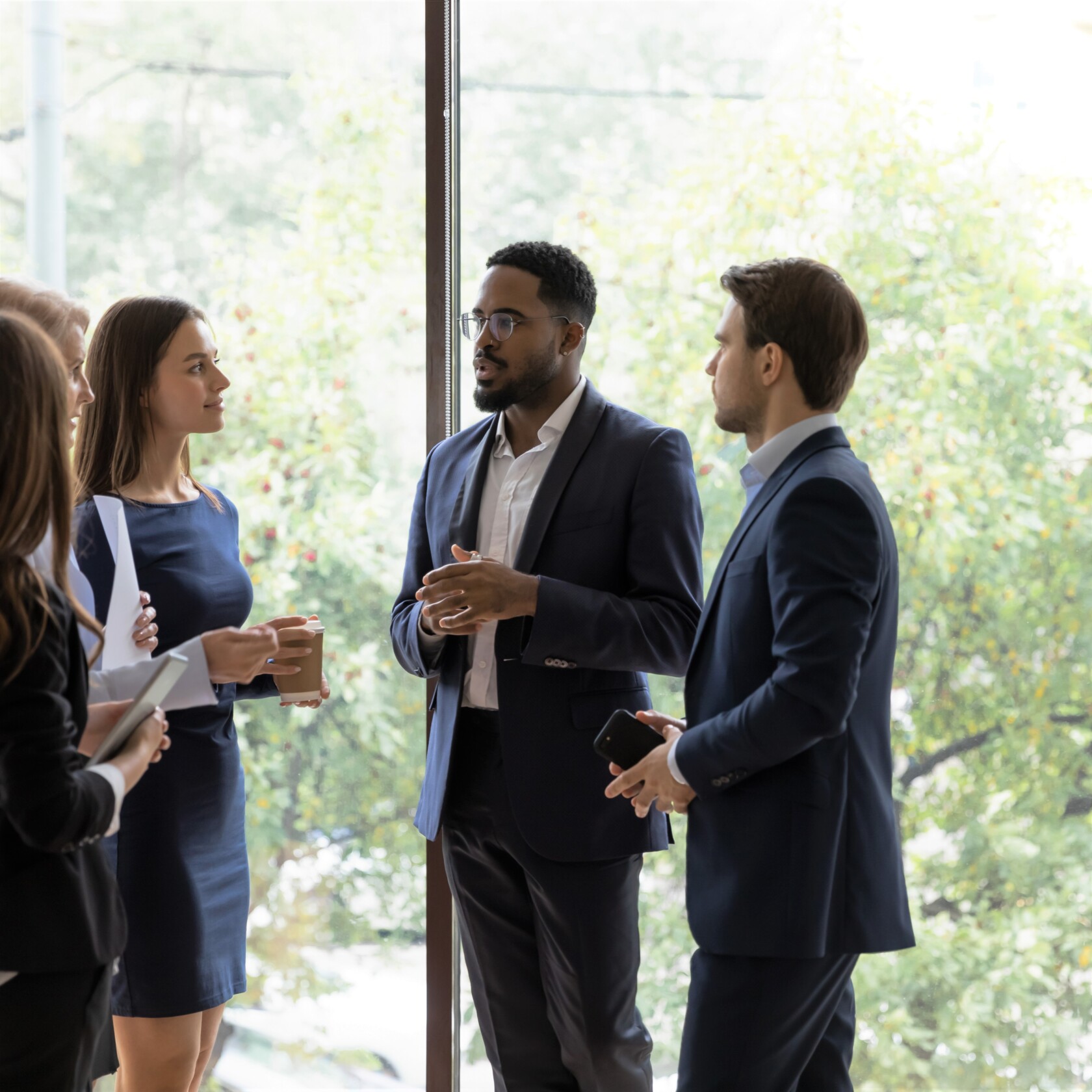 Multiracial colleagues stand in office hallway drink coffee talk brainstorm at work break together. Multiethnic businesspeople employees speak chat discuss ideas or project.