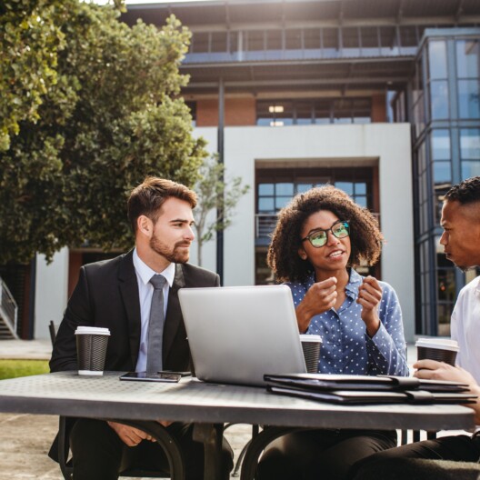 Female executive explaining new business strategy to team while sitting outdoors at table. Business professionals discussing work at office cafeteria.