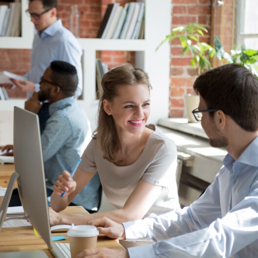 Friendly happy male and female colleagues enjoy fun conversation cooperating in office talking, smiling employees coworkers chatting at workplace have discussion help working together with computer.